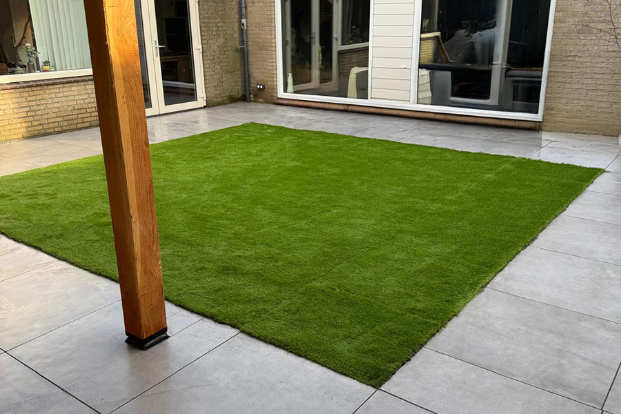Achtertuin met gazon, lichte terrastegels en een houten overkappingspaal